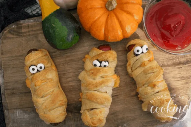 3 mummy hot dogs on a board with eyes and pumpkin gourds and bowl of ketchup