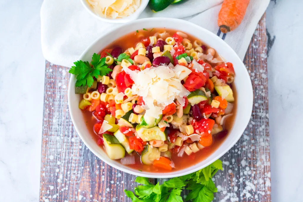 Minestrone soup in white bowl with parsley garnish and grated parmesan cheese.
