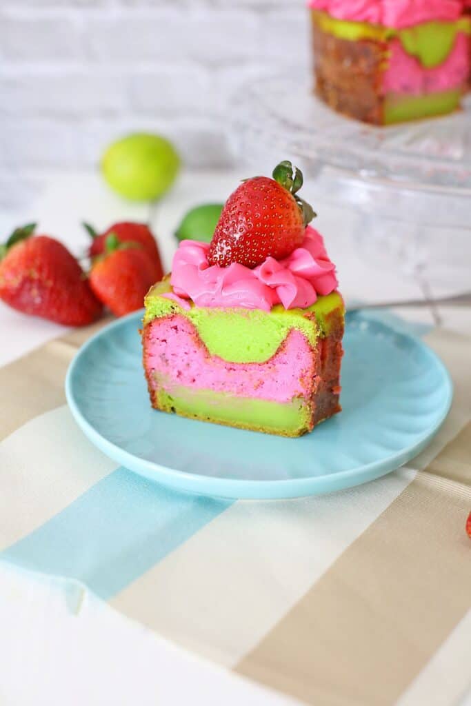 slice of pink and green key lime strawberry cheesecake cake served on a blue plate with strawberries on the side.