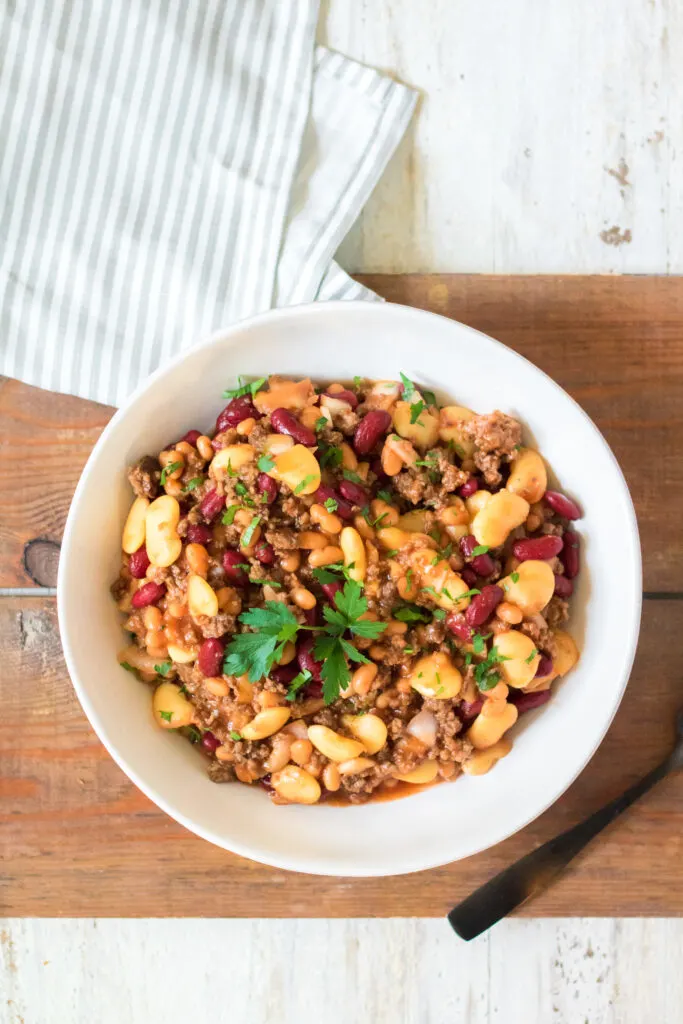 Slow cooker beefy baked beans cooked and served in a white bowl with a pin striped blue napkin.