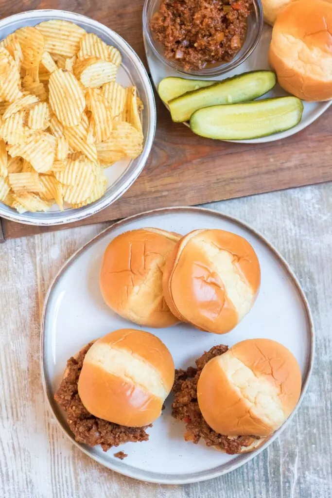 Sloppy Joes sandwiches are served on buns on a white platter with sides of potato chips and pickles