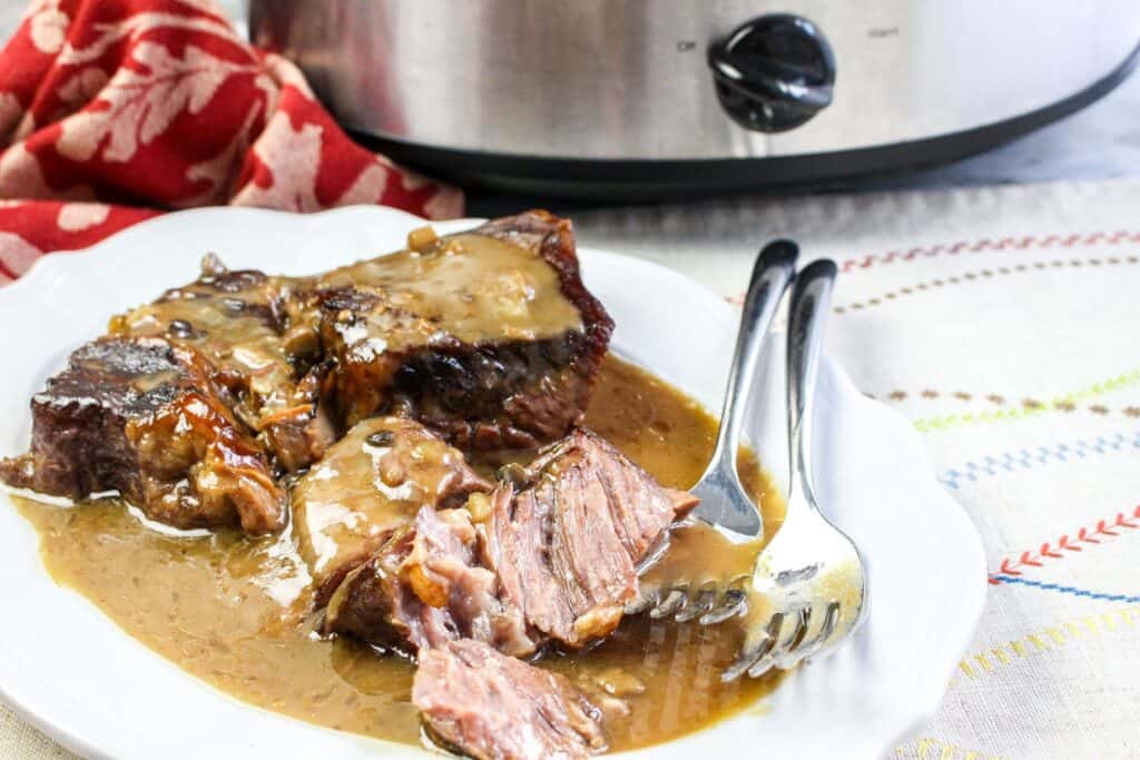 slow cooker roast beef and gravy  vertical served platter with forks crock pot and red towel over a white cloth