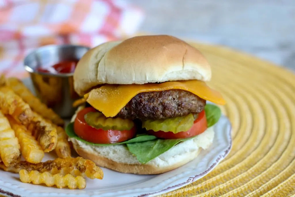 Vertical shot of Air Fryer Burgers with French fries ketchup burger on bun with cheese pickles tomatoes served on white plate with yellow placemat red and white checked towel in background