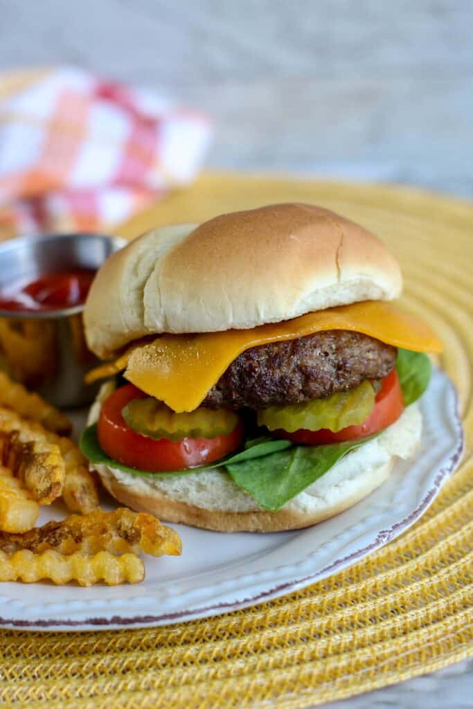 vertical shot close up air fryer burger with cheese pickles tomato lettuce on a bun french fries and ketchup on plate over placemat with checked towels