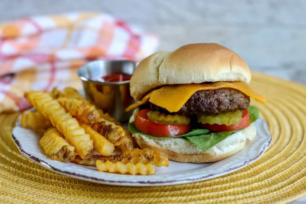 Horizontal air fryer burger with cheese pickles tomato lettuce on a bun french fries and ketchup on plate over placemat with checked towels