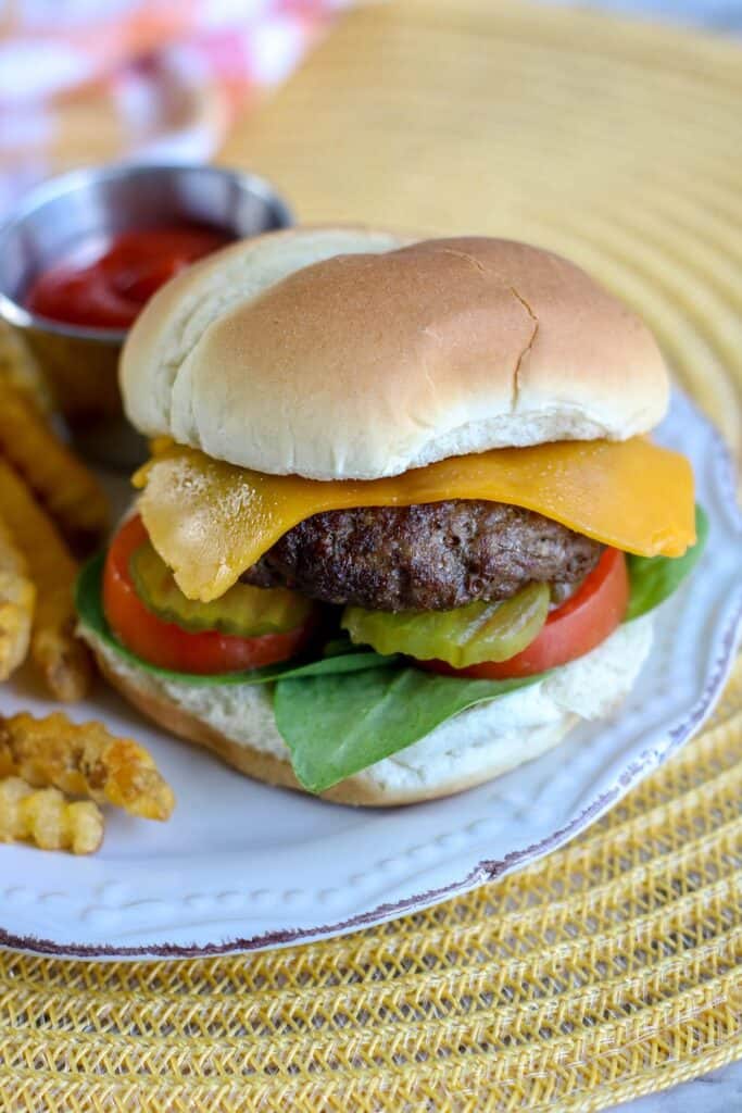 Top shot of Air Fryer Burgers with French fries ketchup burger on bun with cheese pickles tomatoes served on white plate with yellow placemat red and white checked towel in background