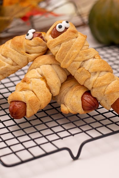 4 Mummy hot dogs with eyes stacked on a cooling rack