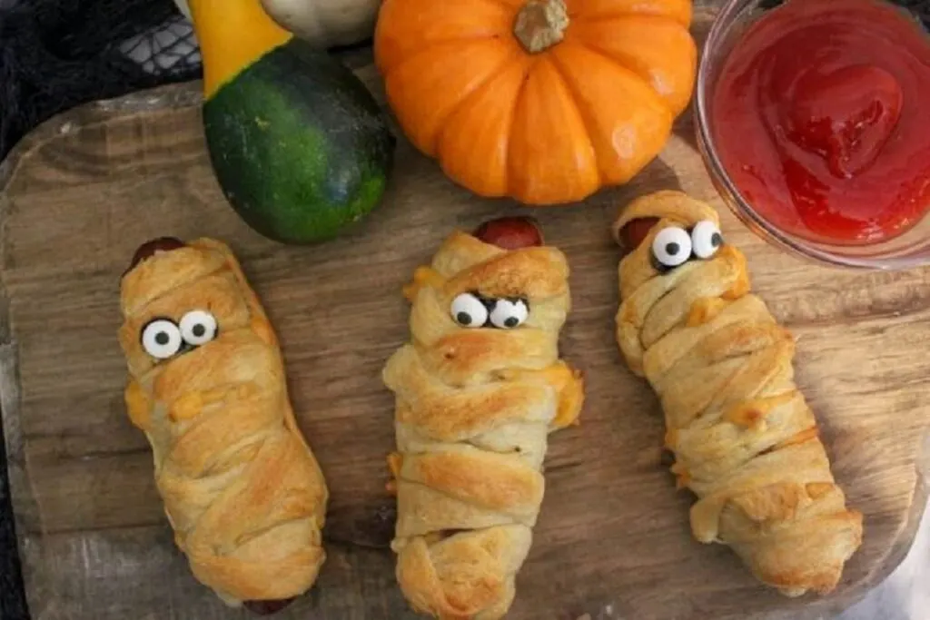 3 Mummy Hot dogs on wooden board with ketchup and festive gourd