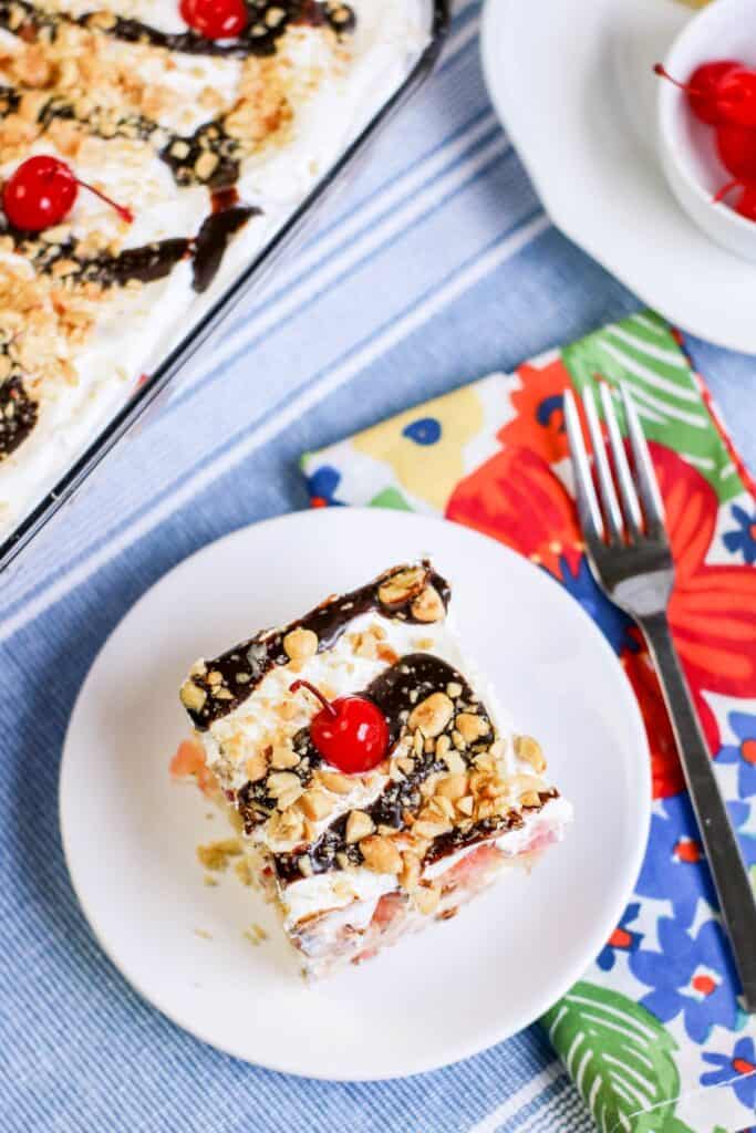 no-bake banana split cake ready to serve on white plate napkin fork cherries and full dessert in a pyrex dish in background
