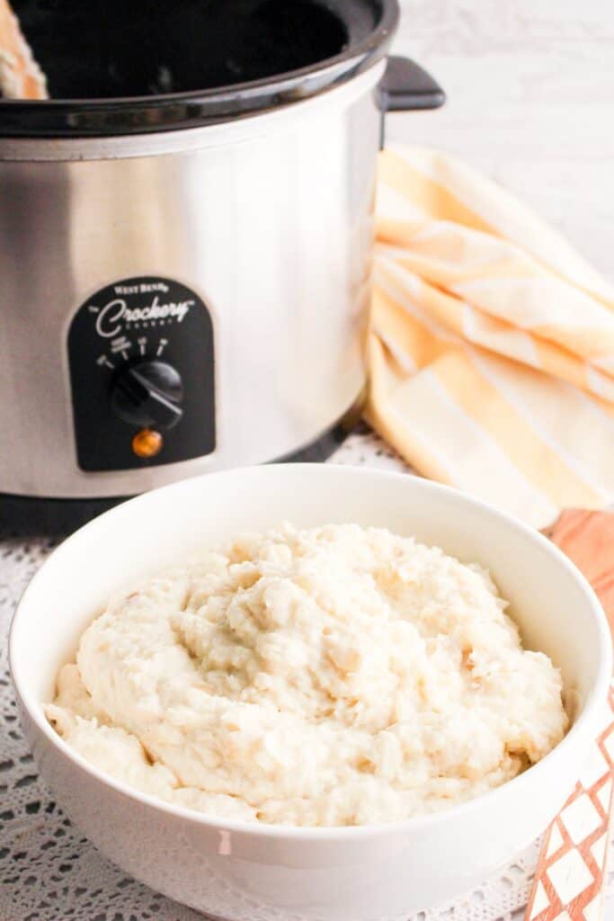 Bowl of mashed potatoes served over lace table cloth with slow cooker and yellow striped towel
