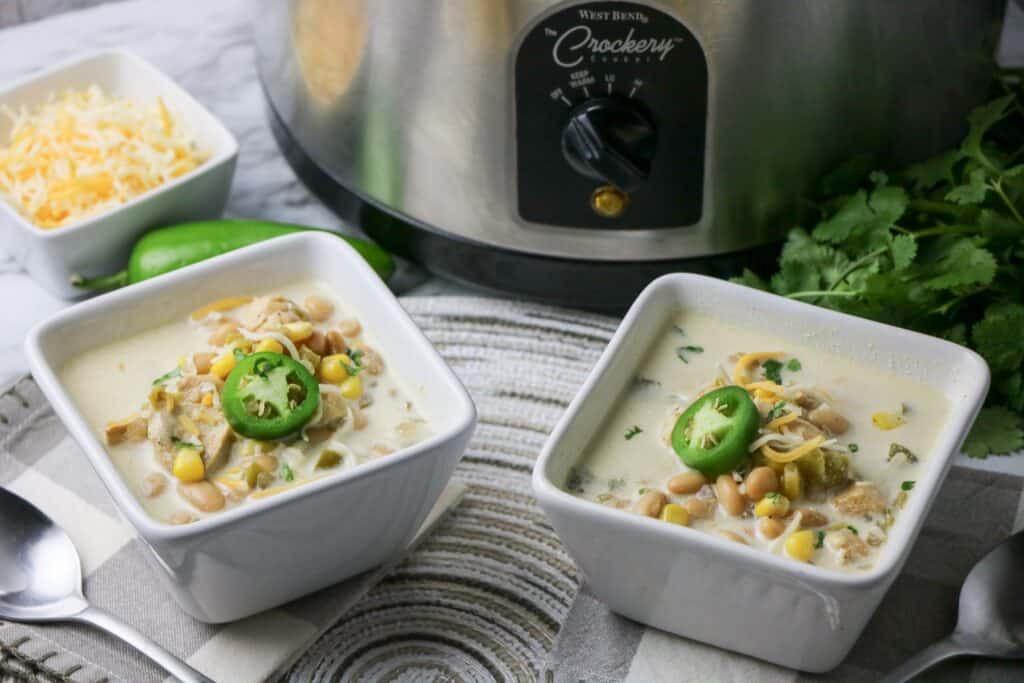 2 bowls Slow Cooker White Bean and Chicken Enchilada Soup in white square bowls with jalapeno pepper slice on top whole jalapeno pepper cheese garnish and slow cooker gray and white napkin place mat other bowl of soup cilantro slow cooker
