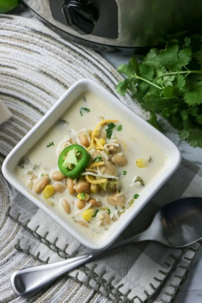 Slow Cooker White Bean and Chicken Enchilada Soup in white square bowl with jalapeno pepper slice on top whole jalapeno pepper cheese and slow cooker cilantro table mat spoon