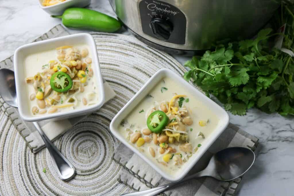  2 bowls with napkins and spoons Slow Cooker White Bean and Chicken Enchilada Soup in white square bowl with jalapeno pepper slice on top whole jalapeno pepper cheese and slow cooker gray and white napkin place mat other bowl of soup cilantro slow cooker