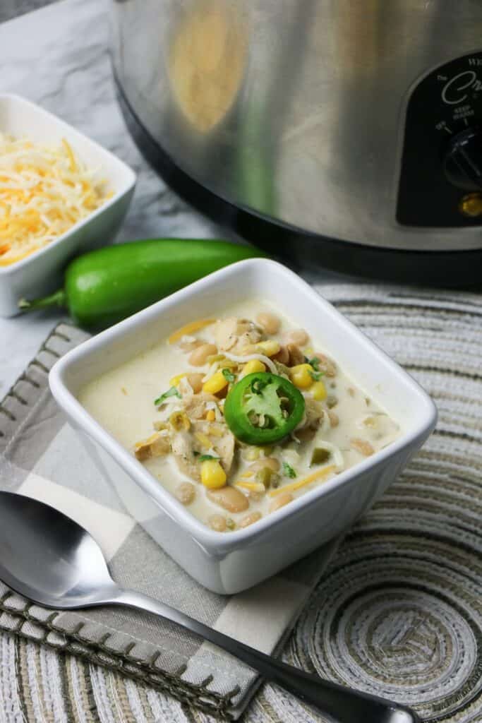 Slow Cooker White Bean and Chicken Enchilada Soup in white square bowl with jalapeno pepper slice on top whole jalapeno pepper cheese and slow cooker