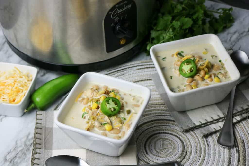 2 bowls of Slow Cooker White Bean and Chicken Enchilada Soup in white square bowl with jalapeno pepper slice on top whole jalapeno pepper cheese garnish and slow cooker gray and white napkin place mat other bowl of soup cilantro slow cooker