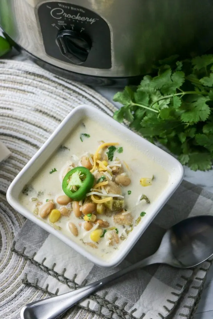 Slow Cooker White Bean and Chicken Enchilada Soup in white square bowl with jalapeno pepper slice on top whole jalapeno pepper cheese and slow cooker gray and white napkin place mat cilantro slow cooker