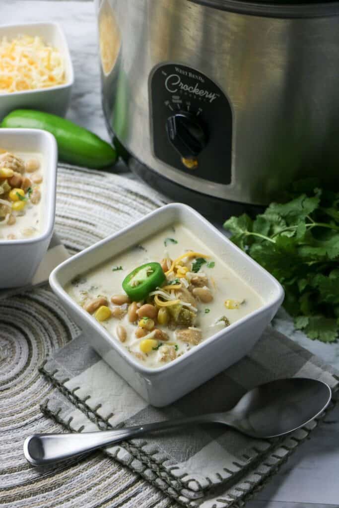Slow Cooker White Bean and Chicken Enchilada Soup in white square bowl with jalapeno pepper slice on top whole jalapeno pepper cheese and slow cooker gray and white napkin place mat other bowl of soup cilantro slow cooker