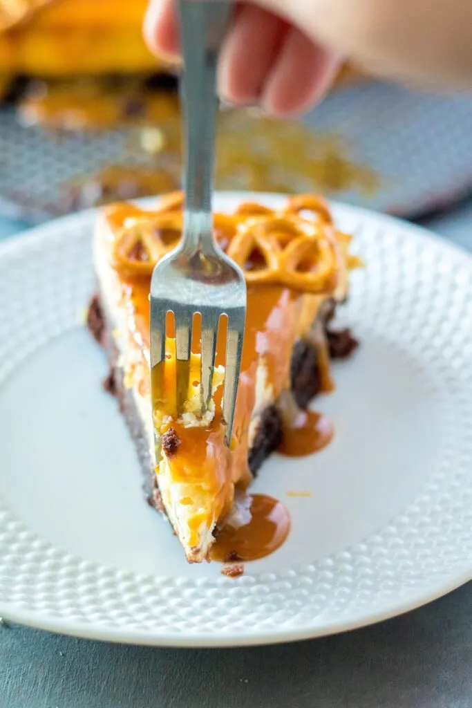 slice of brownie bottom salted caramel cheesecake on white plate cheesecake in back with fork in cheesecake