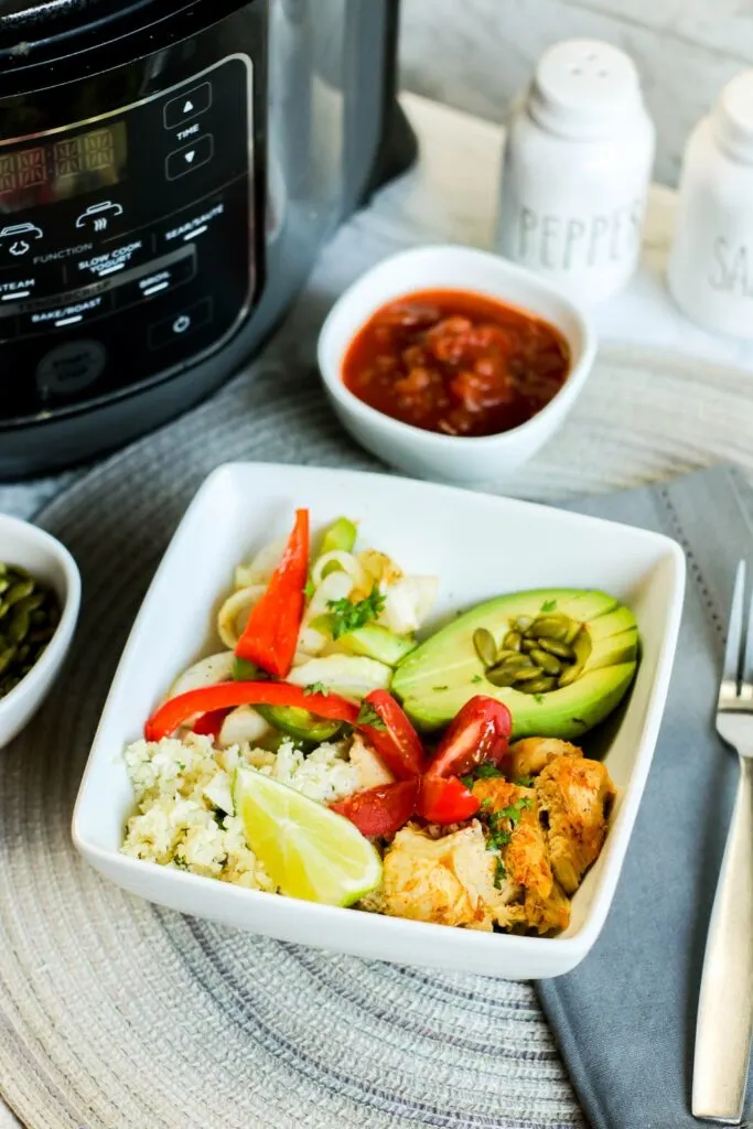 vertical instant pot low carb chicken fajita bowl filled with onions and peppers pepitas salsa avocado, cauli rice tomato on blue table cloth instant pot with fork