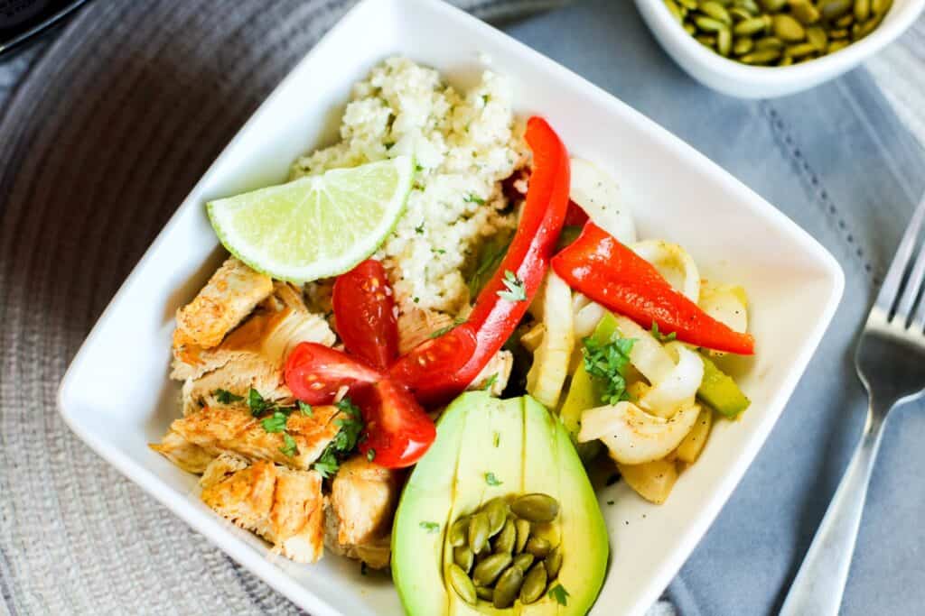 horizontal shot instant pot low carb chicken fajita bowl filled with onions and peppers pepitas salsa avocado, cauli rice tomato on blue table cloth 