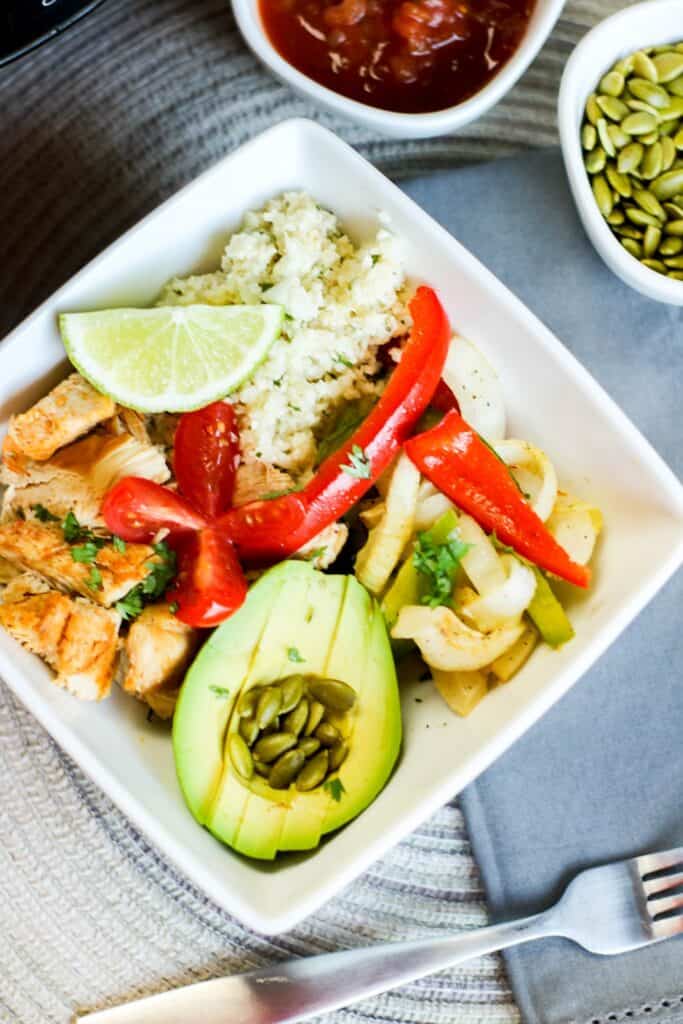 vertical top image instant pot low carb chicken fajita bowl filled with onions and peppers pepitas salsa avocado, cauli rice tomato on blue table cloth instant pot