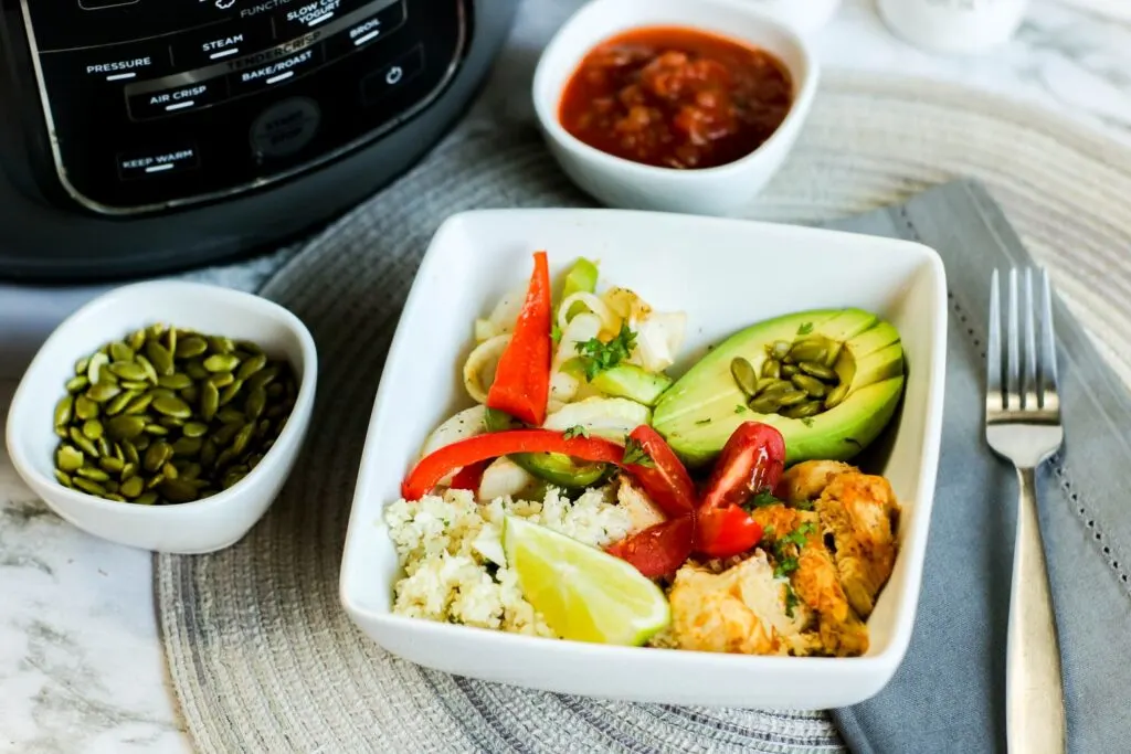 horizontal instant pot low carb sliced chicken fajita bowl filled with onions and peppers pepitas salsa avocado, cauli rice tomato on blue table cloth instant pot fork on blue napkin