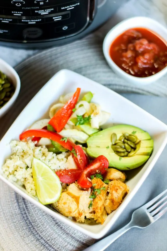 vertical top image instant pot low carb chicken fajita bowl filled with onions and peppers pepitas salsa avocado, cauli rice tomato on blue table cloth instant pot fork