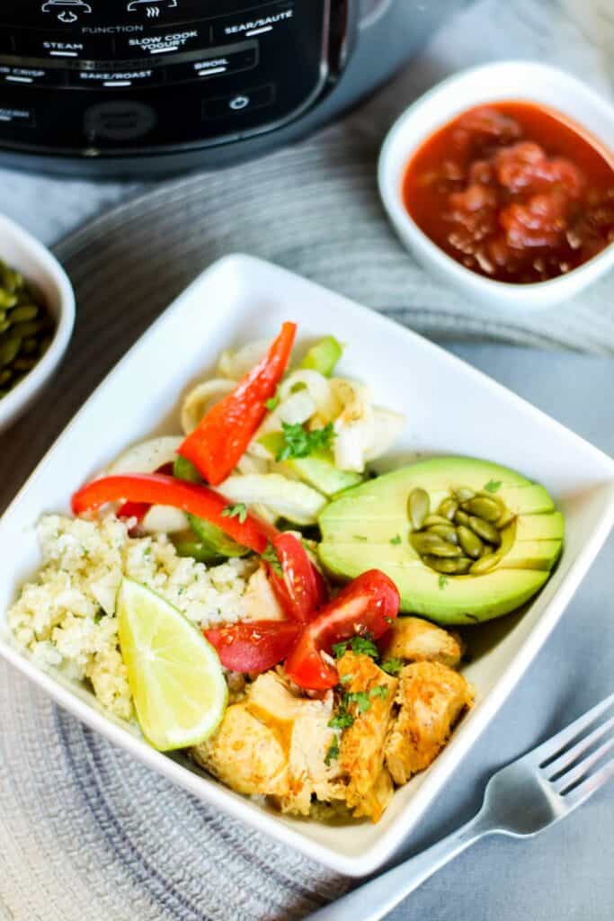 vertical instant pot low carb chicken fajita bowl filled with onions and peppers pepitas salsa avocado, cauli rice tomato on blue table cloth instant pot