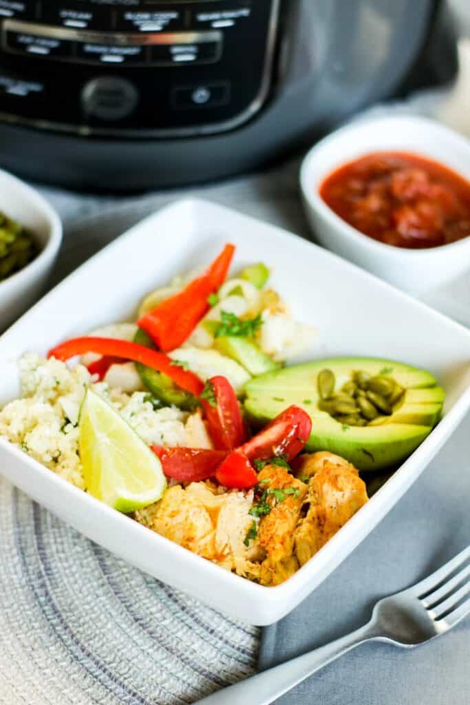 vertical instant pot low carb chicken fajita bowl filled with onions and peppers pepitas salsa avocado, cauli rice tomato on blue table cloth forkinstant pot