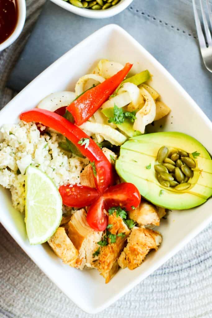 hero instant pot low carb chicken fajita bowl filled with onions and peppers pepitas salsa avocado, cauli rice tomato on blue table cloth instant pot