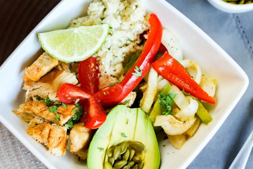 horizontal instant pot low carb chicken pieces fajita bowl filled with onions and peppers pepitas salsa sliced avocado, cauli rice tomato on blue table cloth instant pot