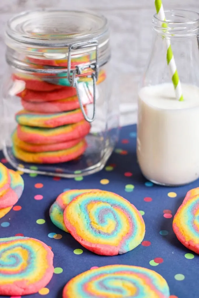 vertical shot rainbow swirl cookies, jar of cookies, jug of milk straw, blue tablecloth polka dots