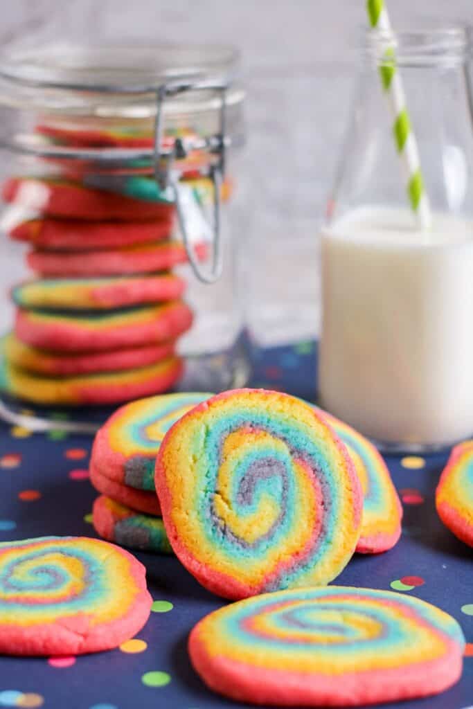 hero vertical shot rainbow swirl cookies, jar of cookies, jug of milk straw, blue tablecloth