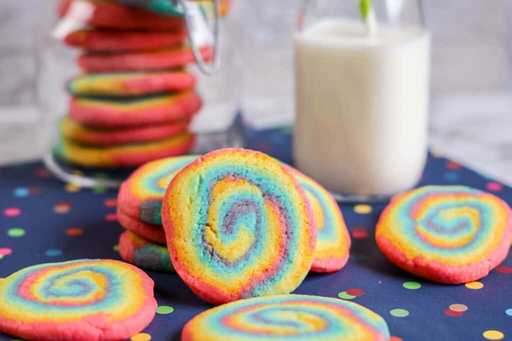 horizontal shot rainbow swirl cookies, jar of cookies, jug of milk straw, blue tablecloth