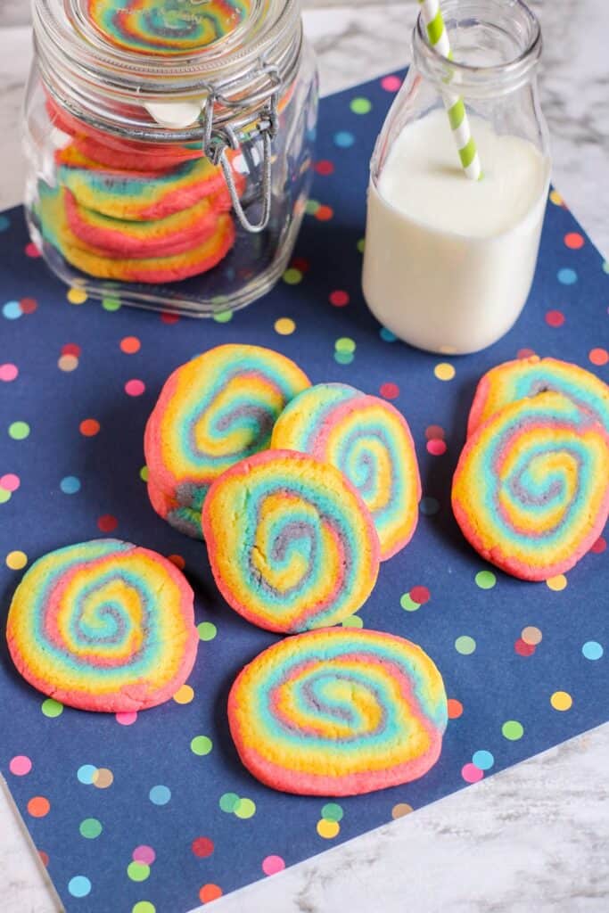 vertical above shot rainbow swirl cookies, jar of cookies, jug of milk straw, blue tablecloth