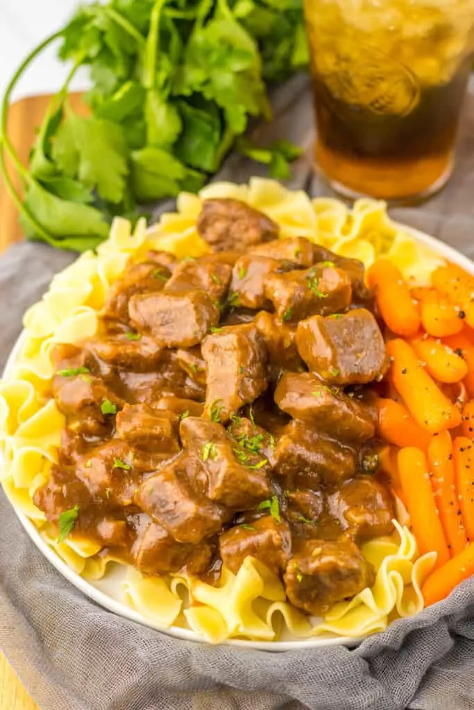 beef tips, carrots over noodles served in white bowl, gray mat arugula and iced tea background