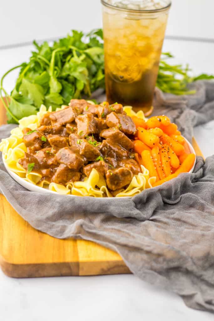 Slow cooker beef tips and gravy served in white bowl over noodles with baby carrots arugula background, iced tea over gray napkin