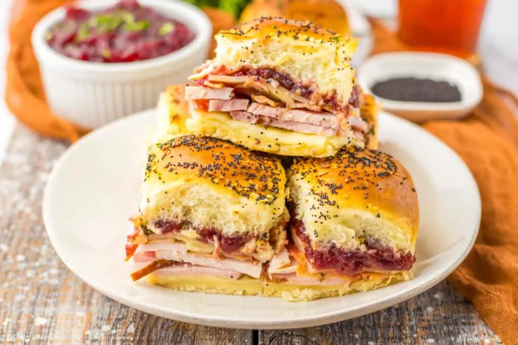 Cranberry Club Sliders stacked for serving with ham, turkey cheese, and fluffy roll top loaded with poppy seeds.