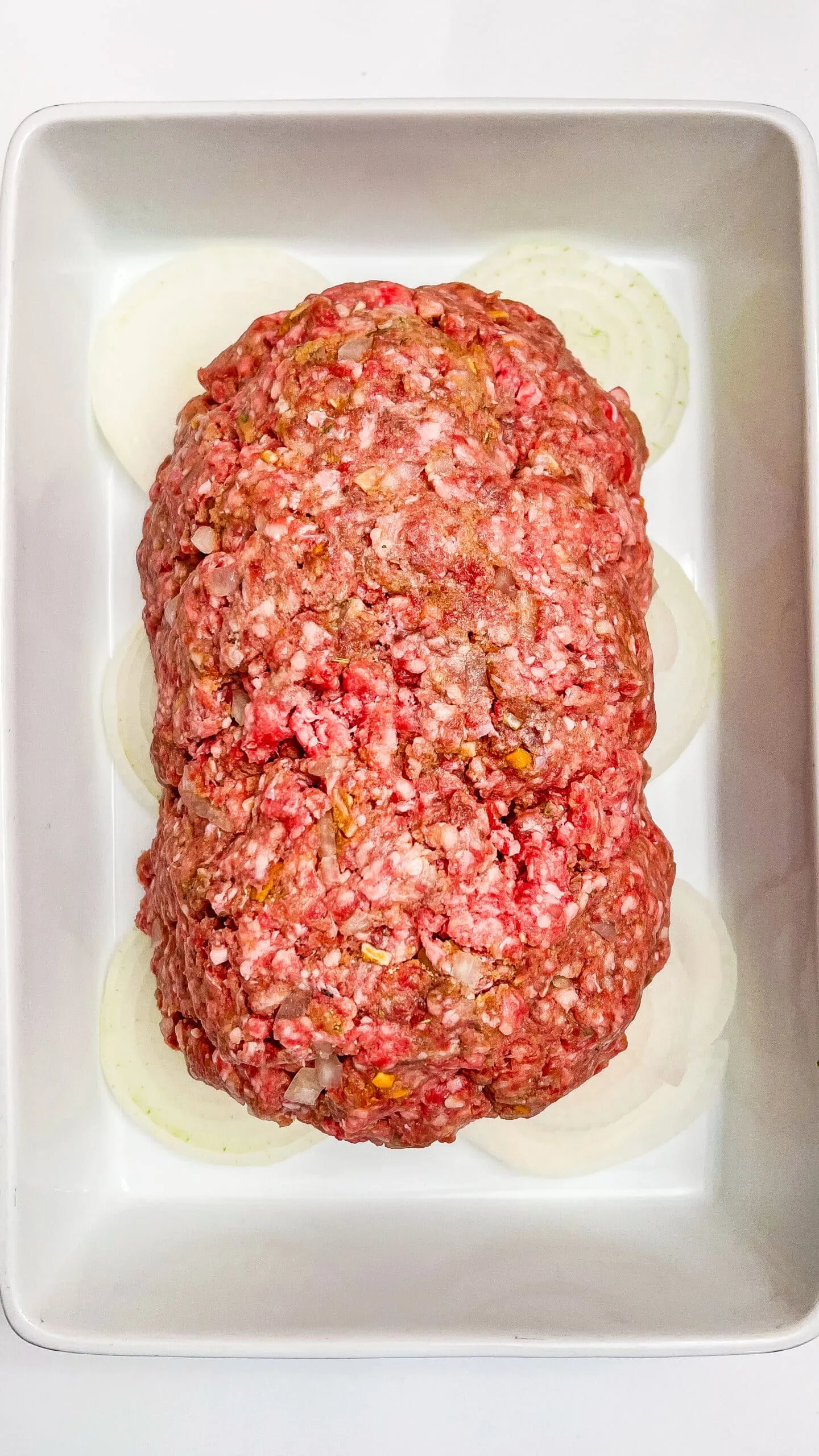 Delicious homemade meatloaf topped with seasoned ground beef, ready for baking with onion slices in dish.