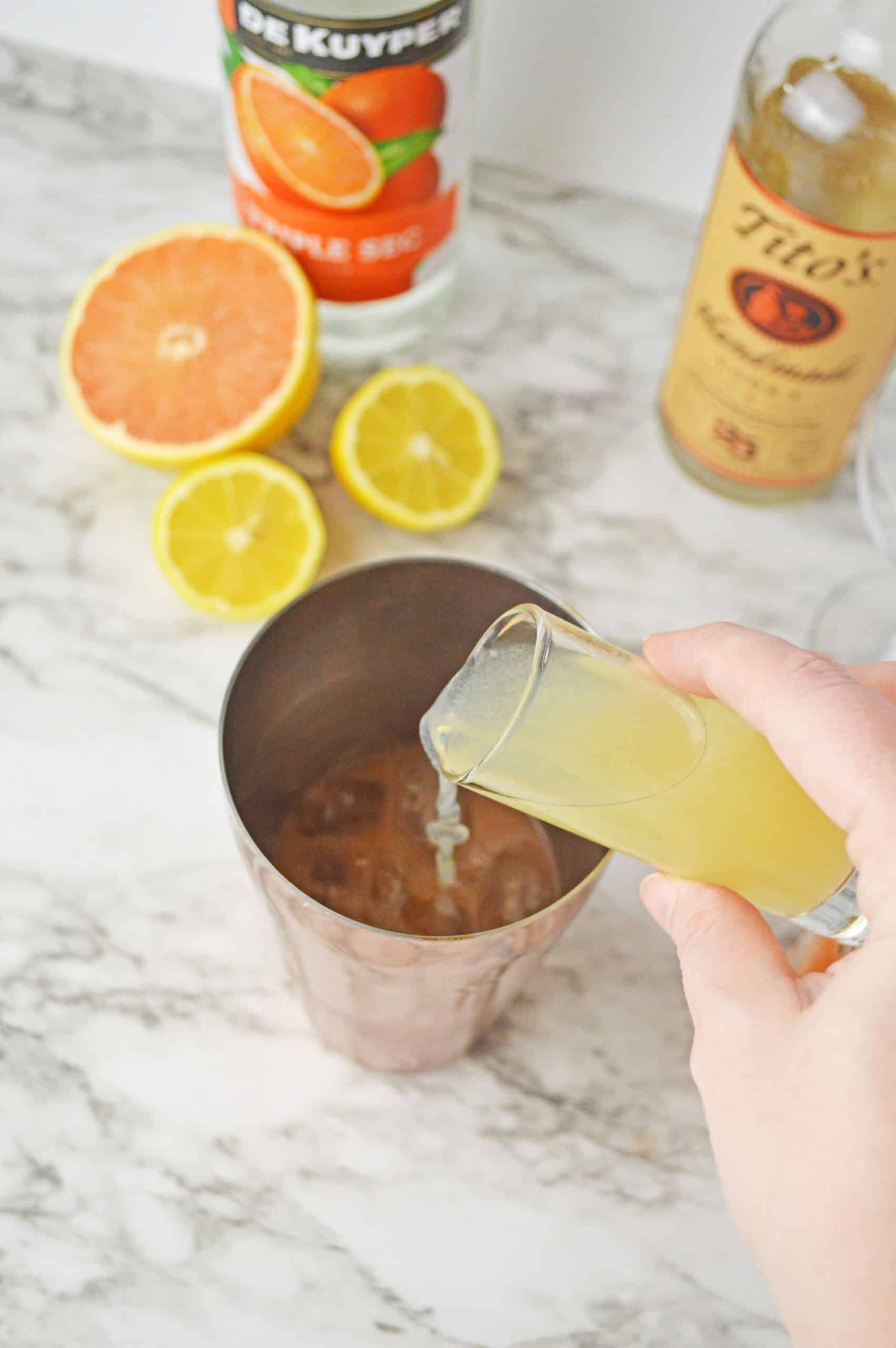 Hand pouring grapefruit cocktail into a copper mug with citrus slices and mixers in the background.
